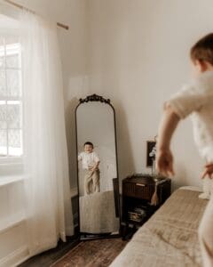 Young boy smiles at his reflection in a vintage mirror during a family session in a softly lit Mendham, NJ studio, designed to feel like home with timeless styling and natural light.