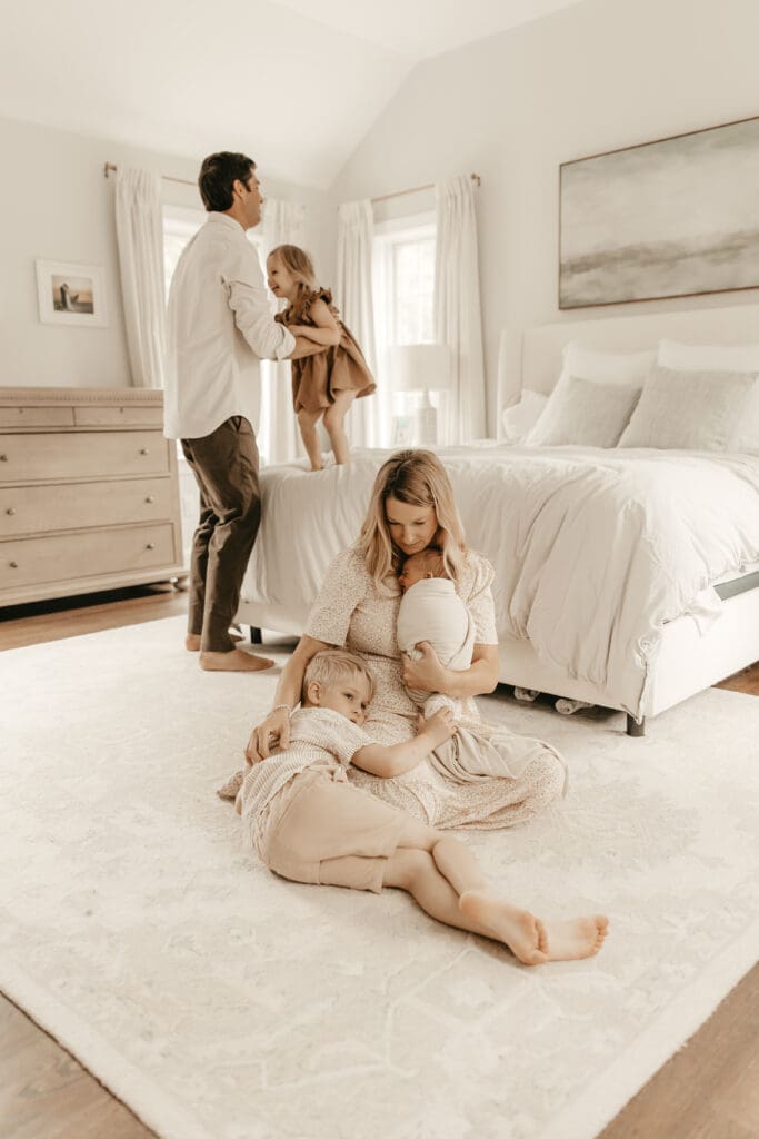 Mother cradling newborn on the bedroom floor while older sibling cuddles close; father lifts toddler onto the bed in a softly lit, neutral-toned room — captured during an in-home newborn and family session in Central New Jersey.