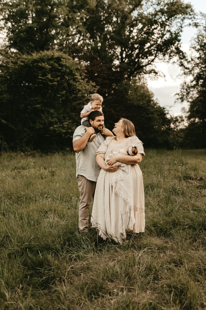 Family of four standing in a lush field at sunset, with dad carrying toddler on shoulders and mom holding newborn — warm, joyful outdoor family session in Mendham, NJ.