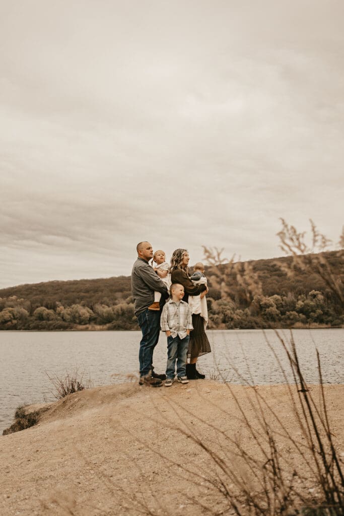 Family of five standing together on a lakeside cliff under a cloudy sky, wrapped in connection with autumn trees in the background — outdoor family photography session in Stanhope, New Jersey.